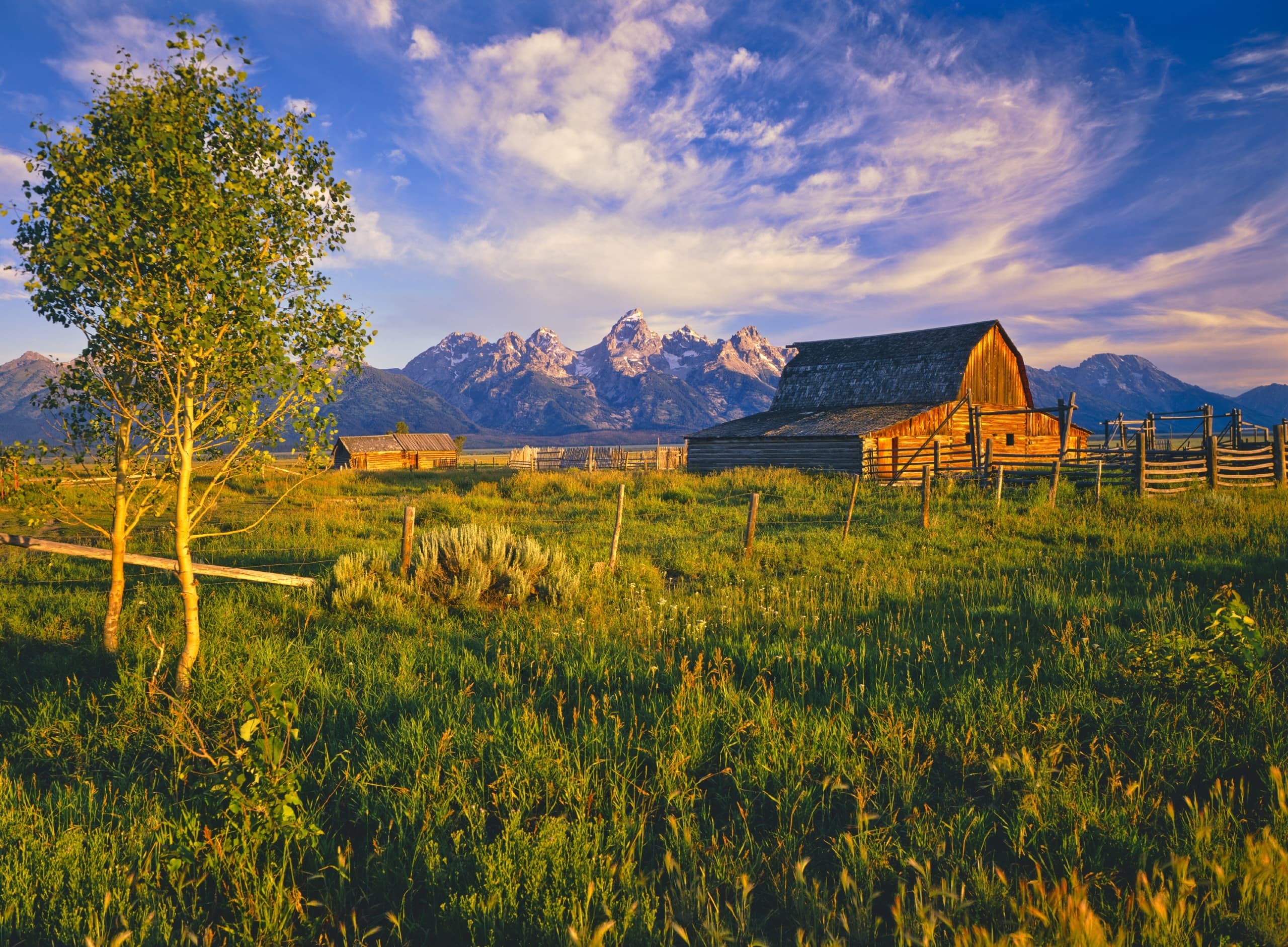 Moulton Barn at sunset in Summer with a view of the surrounding fields and Teton Mountains in the background.